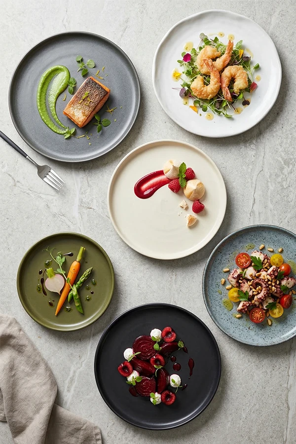 An overhead view of six different gourmet dishes on unique ceramic plates arranged on a grey table.
